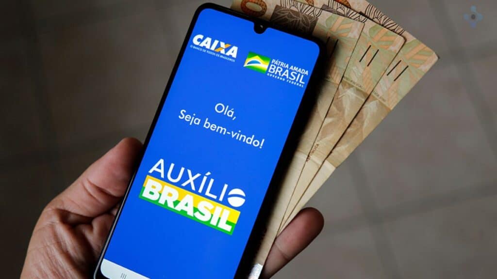 Uma imagem que ilustra uma pessoa segurando um celular com o aplicativo do auxílio Brasil aberto.