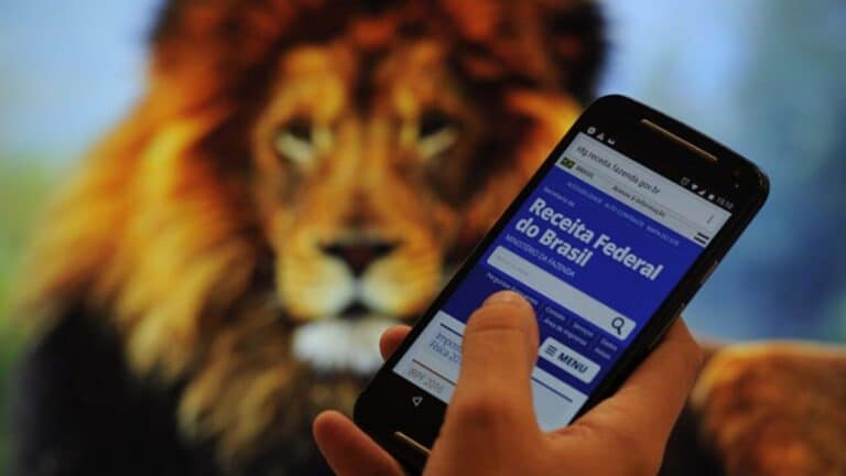 Uma imagem que ilustra uma mão segurando um celular com aplicativo da receita federal aberto e ao fundo uma imagem de leão, que representa o imposto de renda.