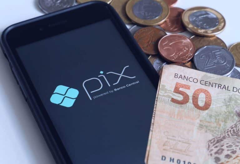 Uma imagem com um celular aberto no aplicativo do PIX e notas e moedas ao redor.